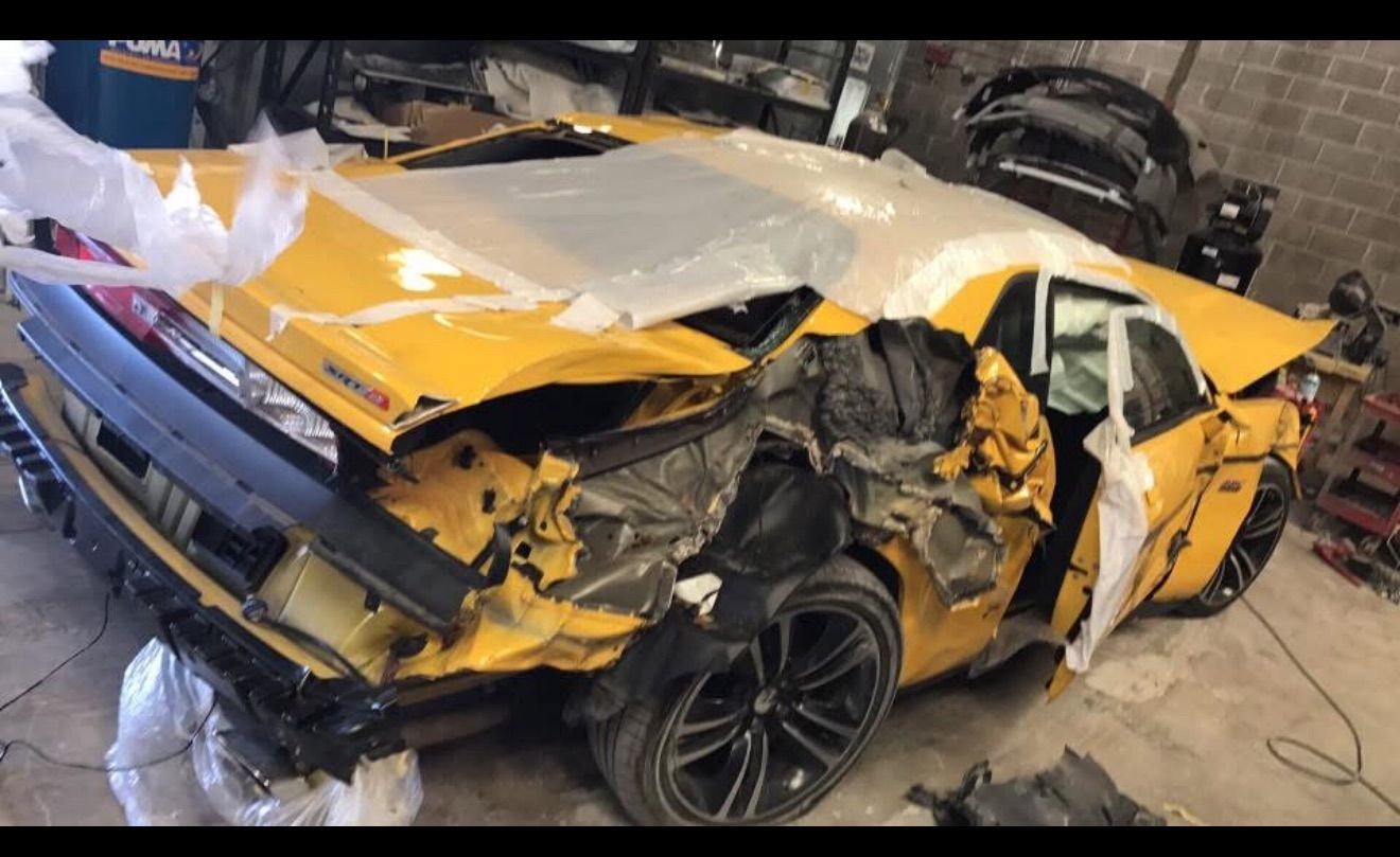 Yellow car severely damaged in what appears to be a collision; front end crumpled, wrapped in plastic, in a repair shop.