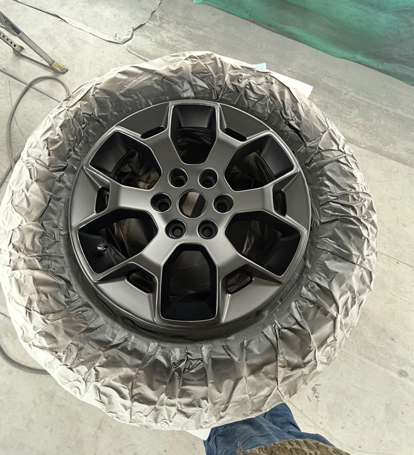 Black car wheel spray-painted, masked tire, gray paint on concrete, person's jeans and boot visible.