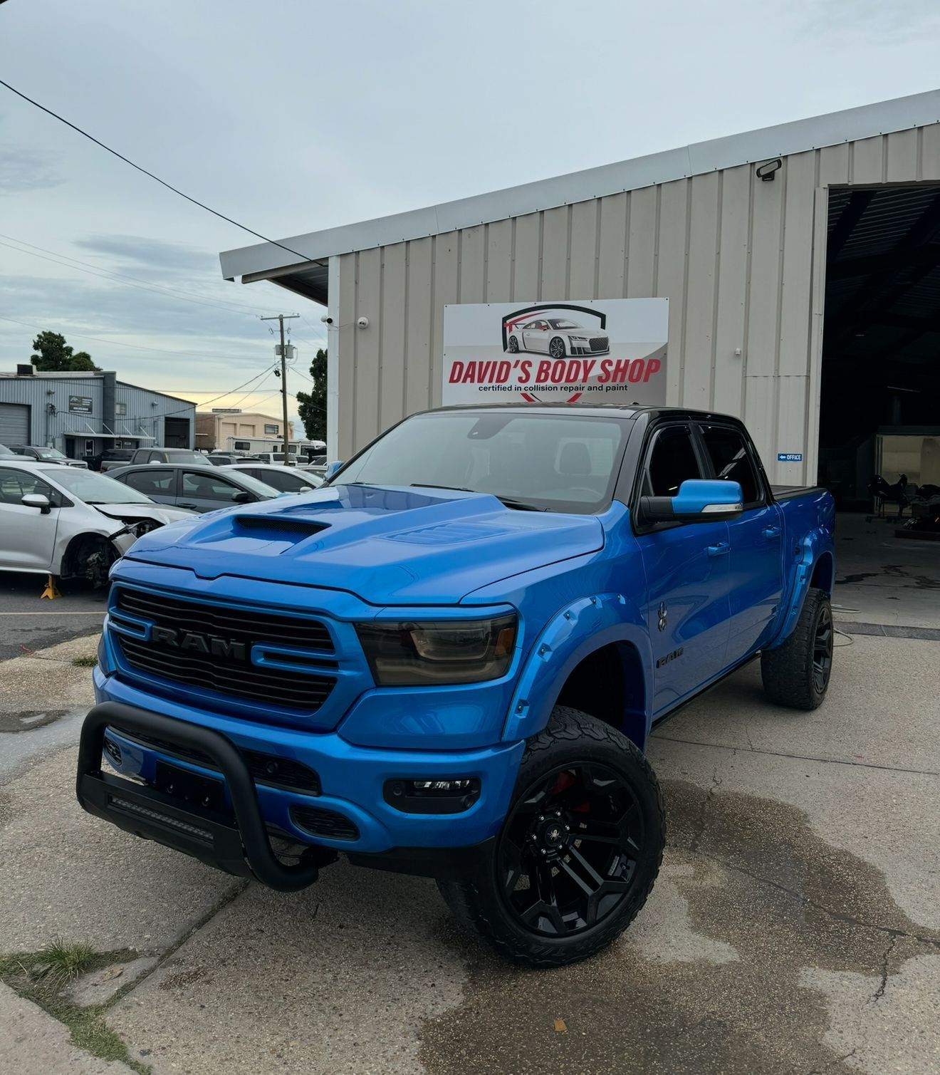 Blue Dodge Ram truck parked outside a body shop with a black bull bar and black wheels.