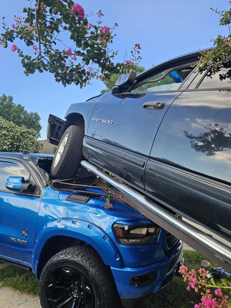 Blue truck crushed under a dark truck. Outdoors, daylight, tree branches above.