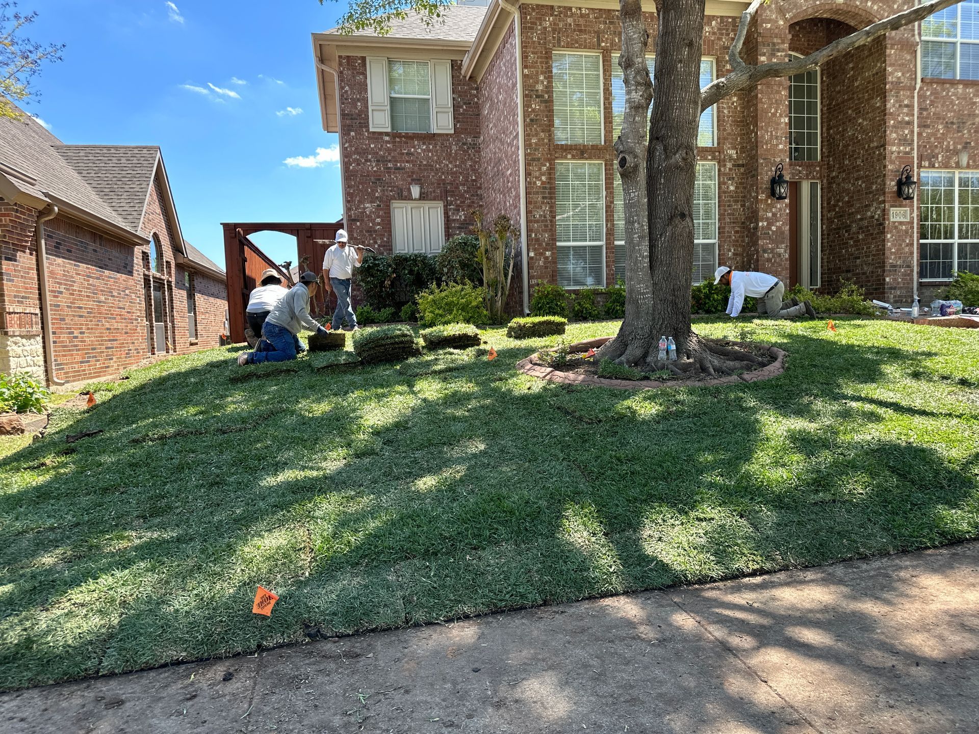 A group of people are working on a lush green lawn in front of a brick house.