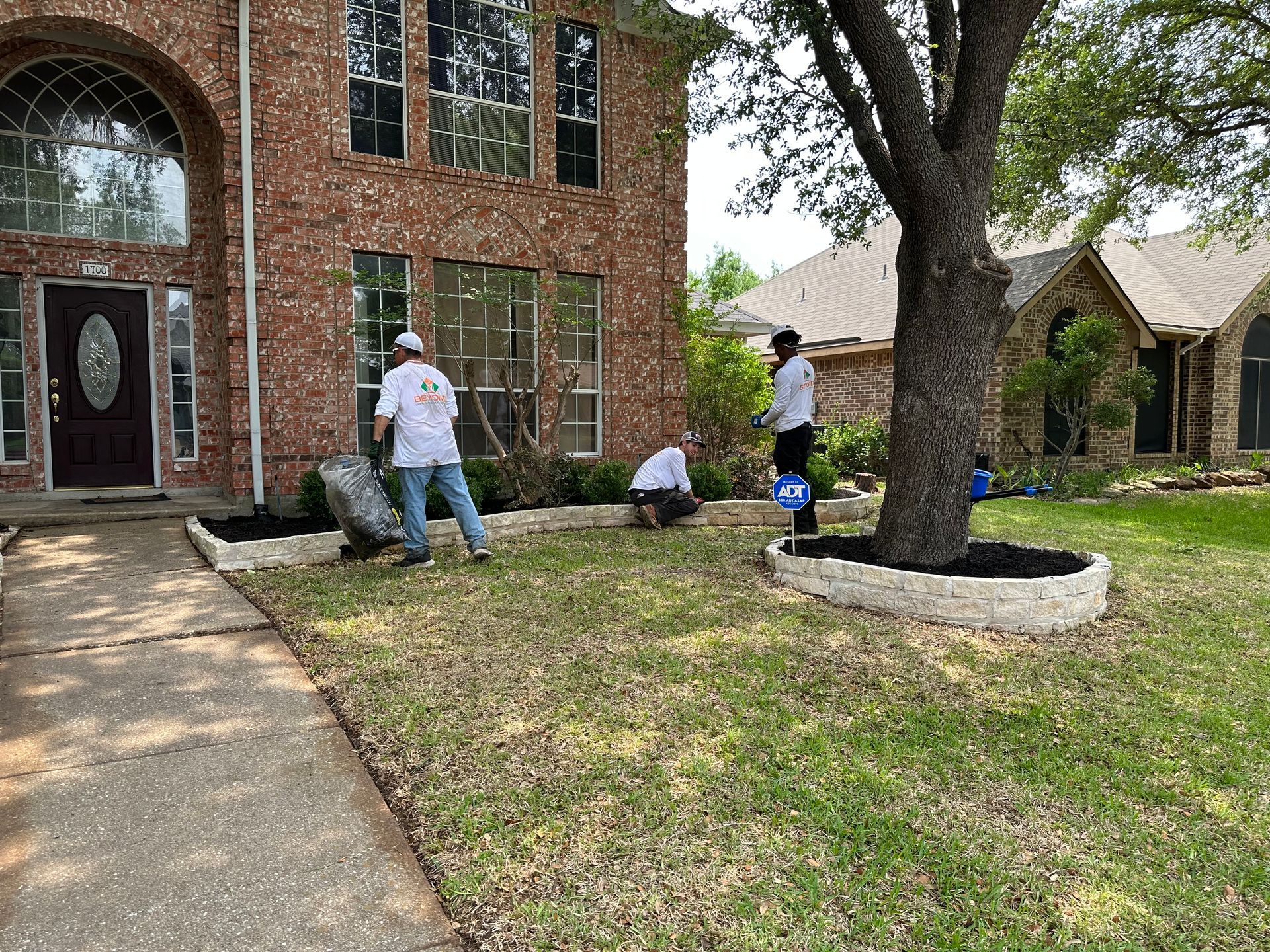 Two men are working on a lawn in front of a large brick house.