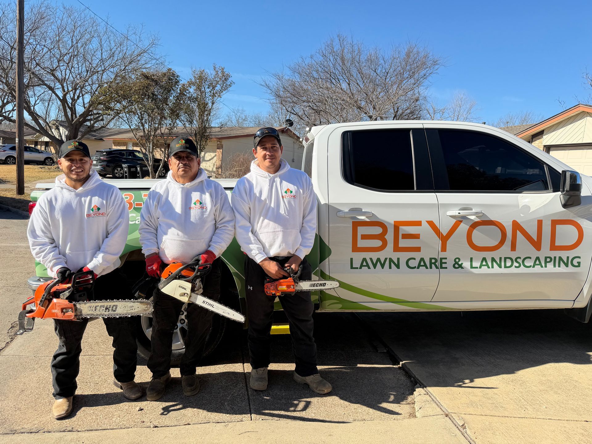 Three workers in white hoodies holding chainsaws stand in front of a white truck branded Beyond Lawn Care & Landscaping.