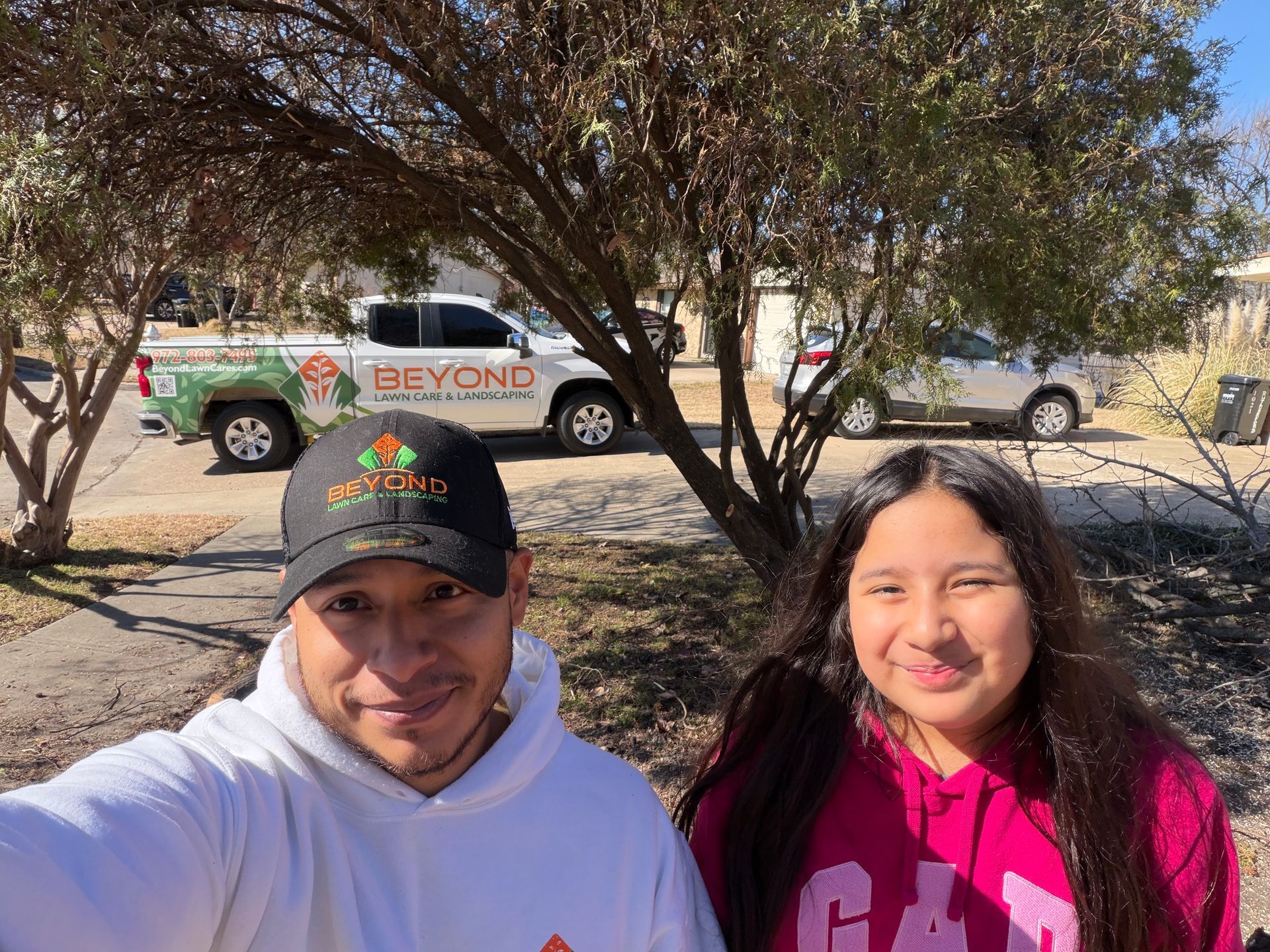 A man wearing a branded baseball cap and hoodie poses for a selfie with a girl outdoors, with service trucks in the back.