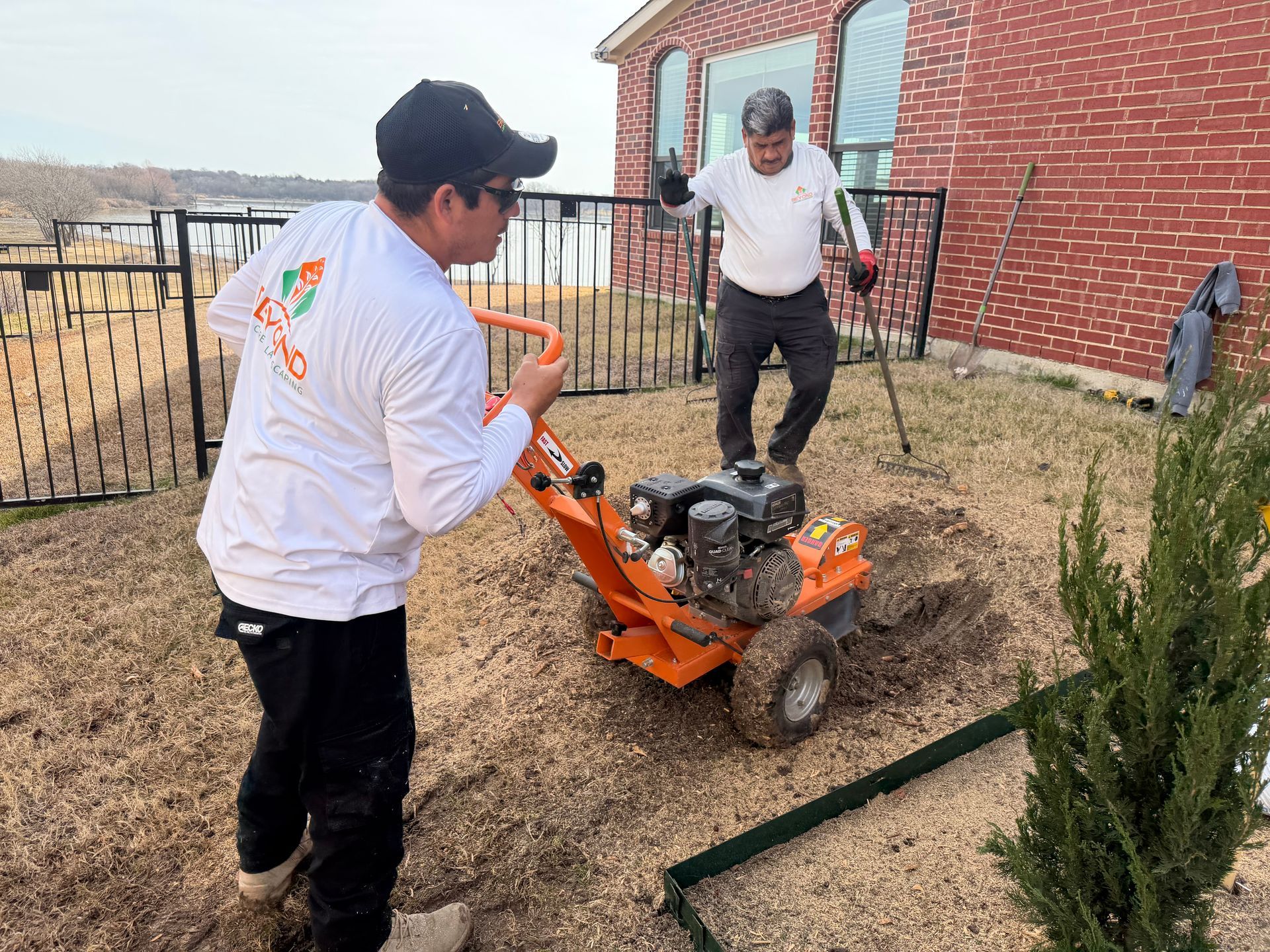 Two workers use an orange gas-powered trencher to dig in a yard next to a red brick house.
