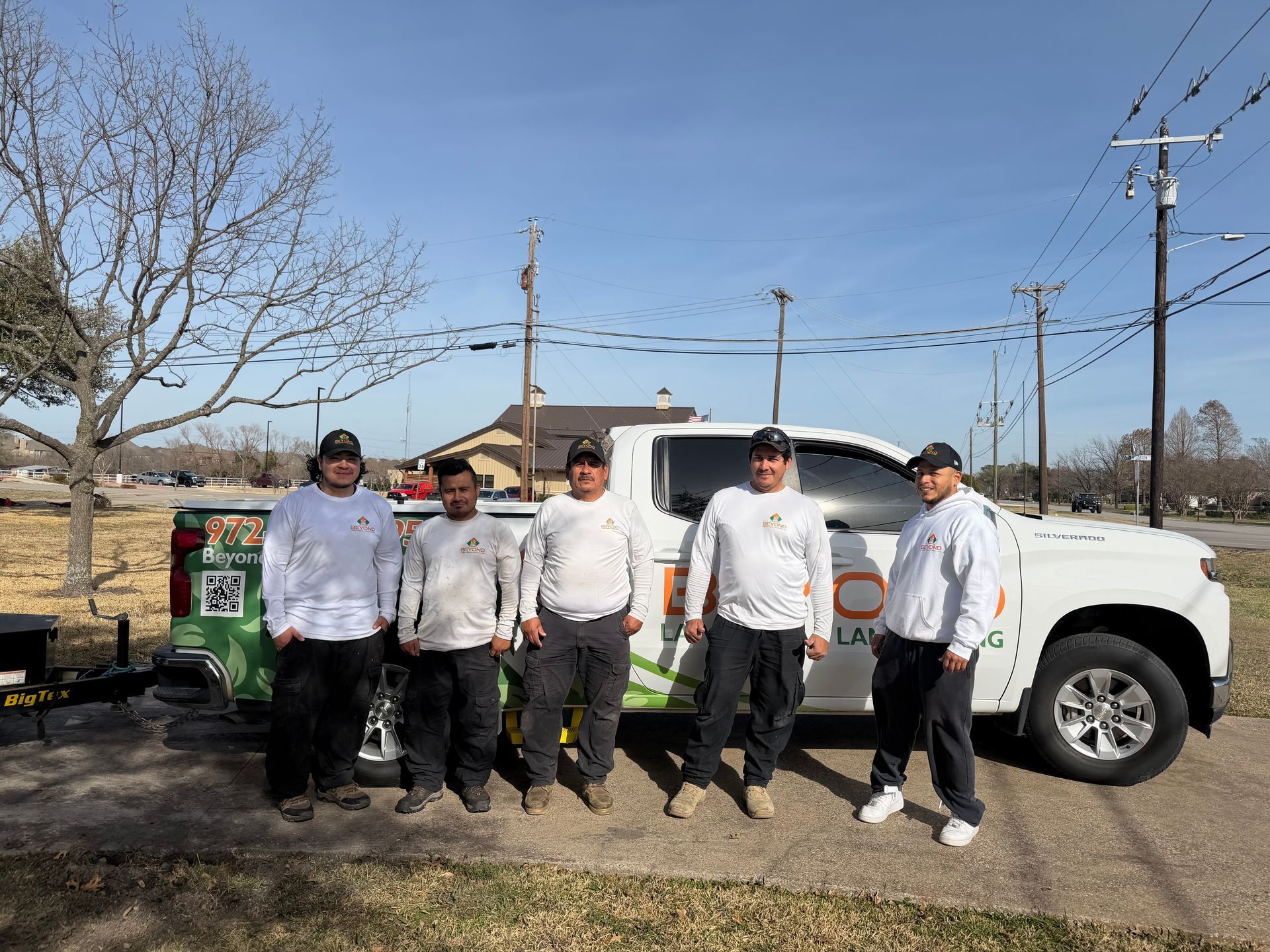 Five workers in white long-sleeved shirts and black pants pose in front of a white work truck parked on a paved lot.