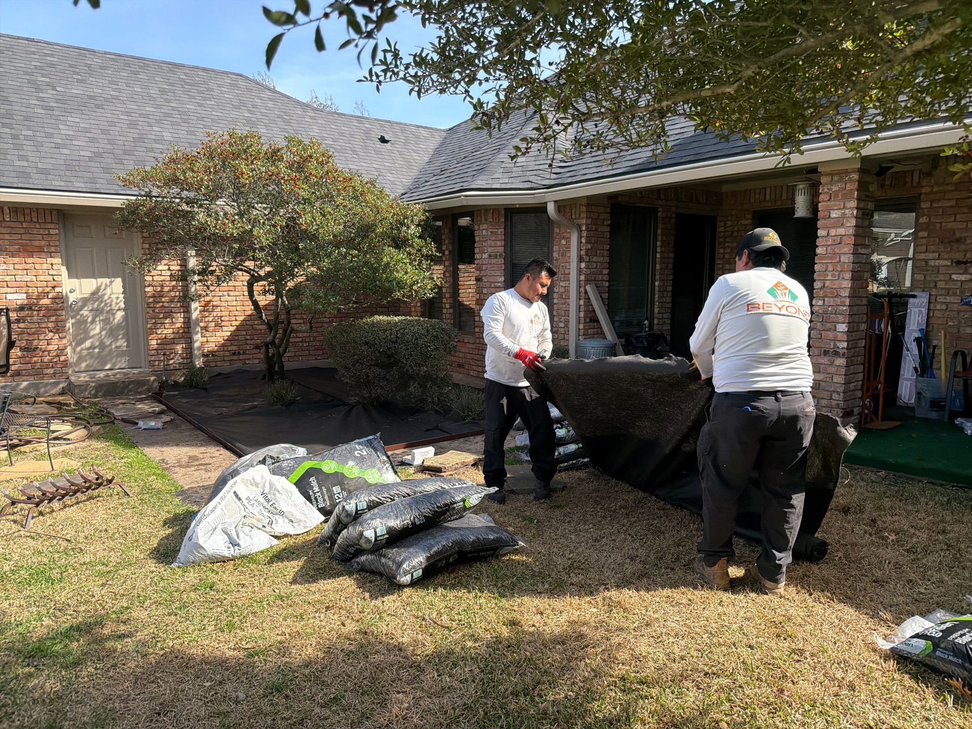 Two workers in long-sleeved shirts roll out a large black landscaping tarp in a residential yard near a brick house.
