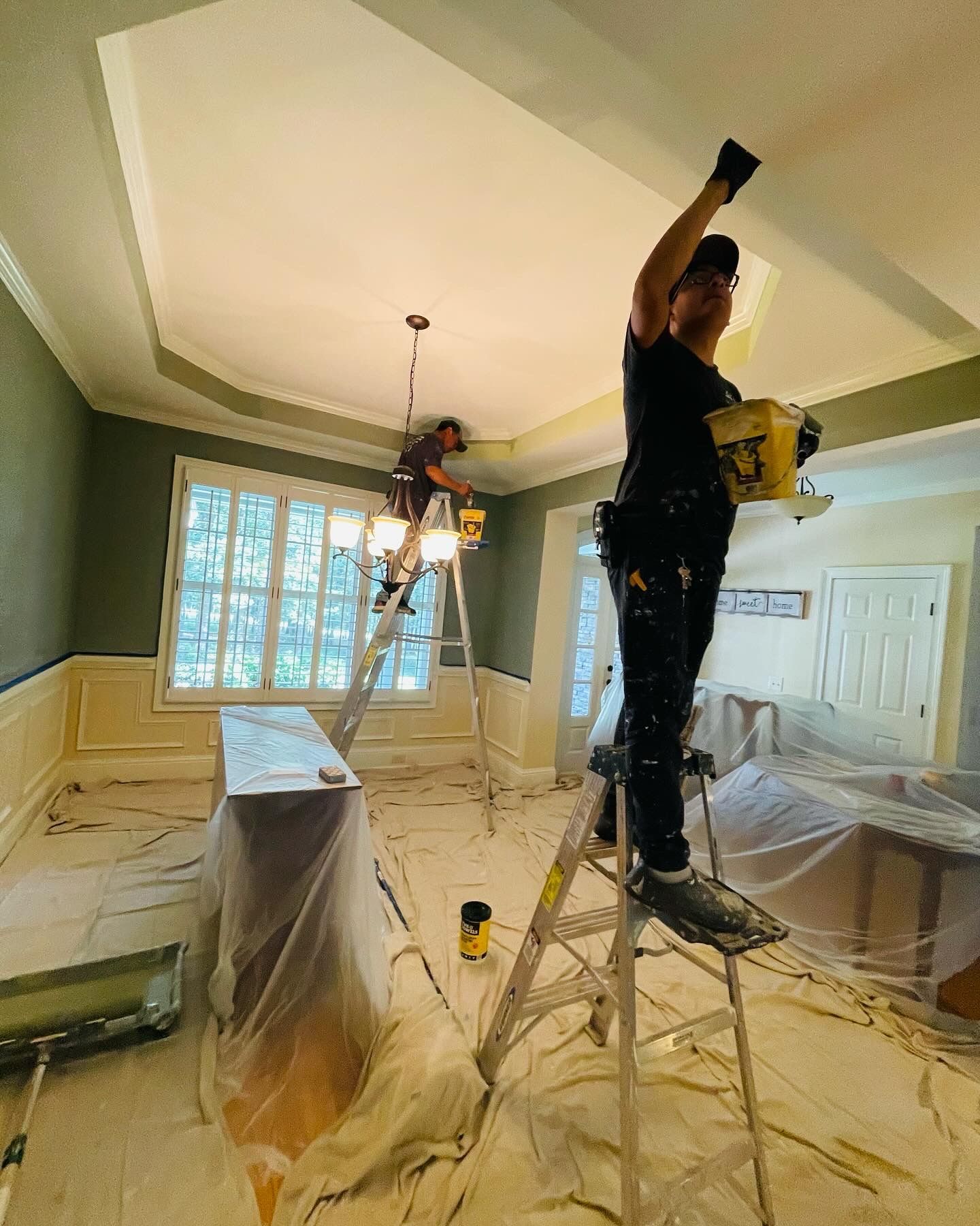A man is standing on a ladder in a living room painting the ceiling.