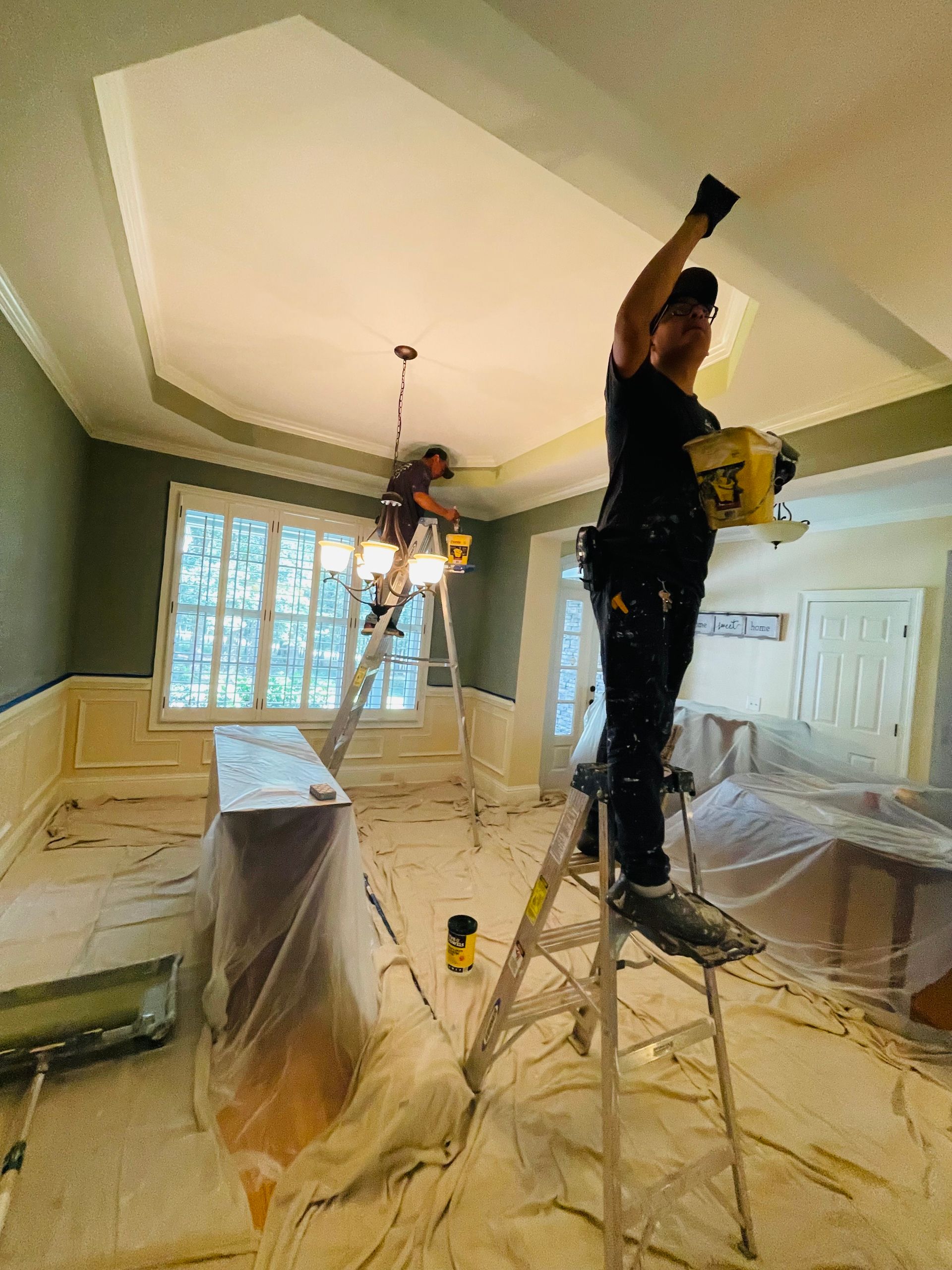 A man is standing on a ladder in a living room painting the ceiling.