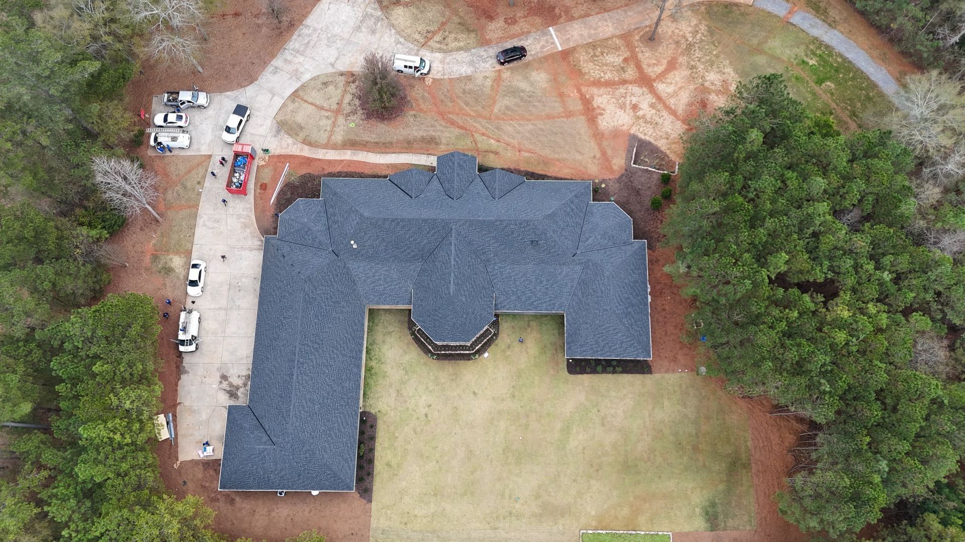 Aerial view of a dark-roofed, L-shaped house with a lawn, driveway, and surrounding trees.