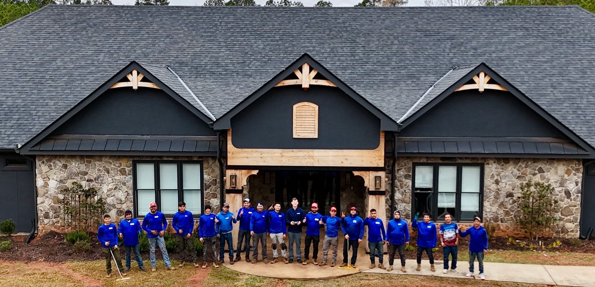 A group of people standing in front of a stone-faced building with three gables under a dark gray roof.