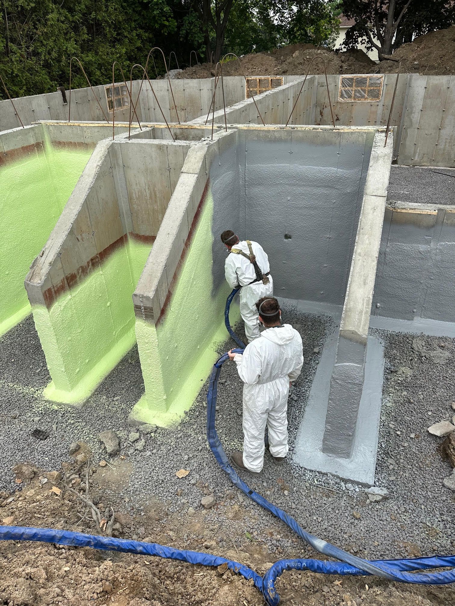 Deux ouvriers en combinaisons blanches projettent de l'isolant sur une fondation en béton. Des sections du mur sont recouvertes d'isolant vert.