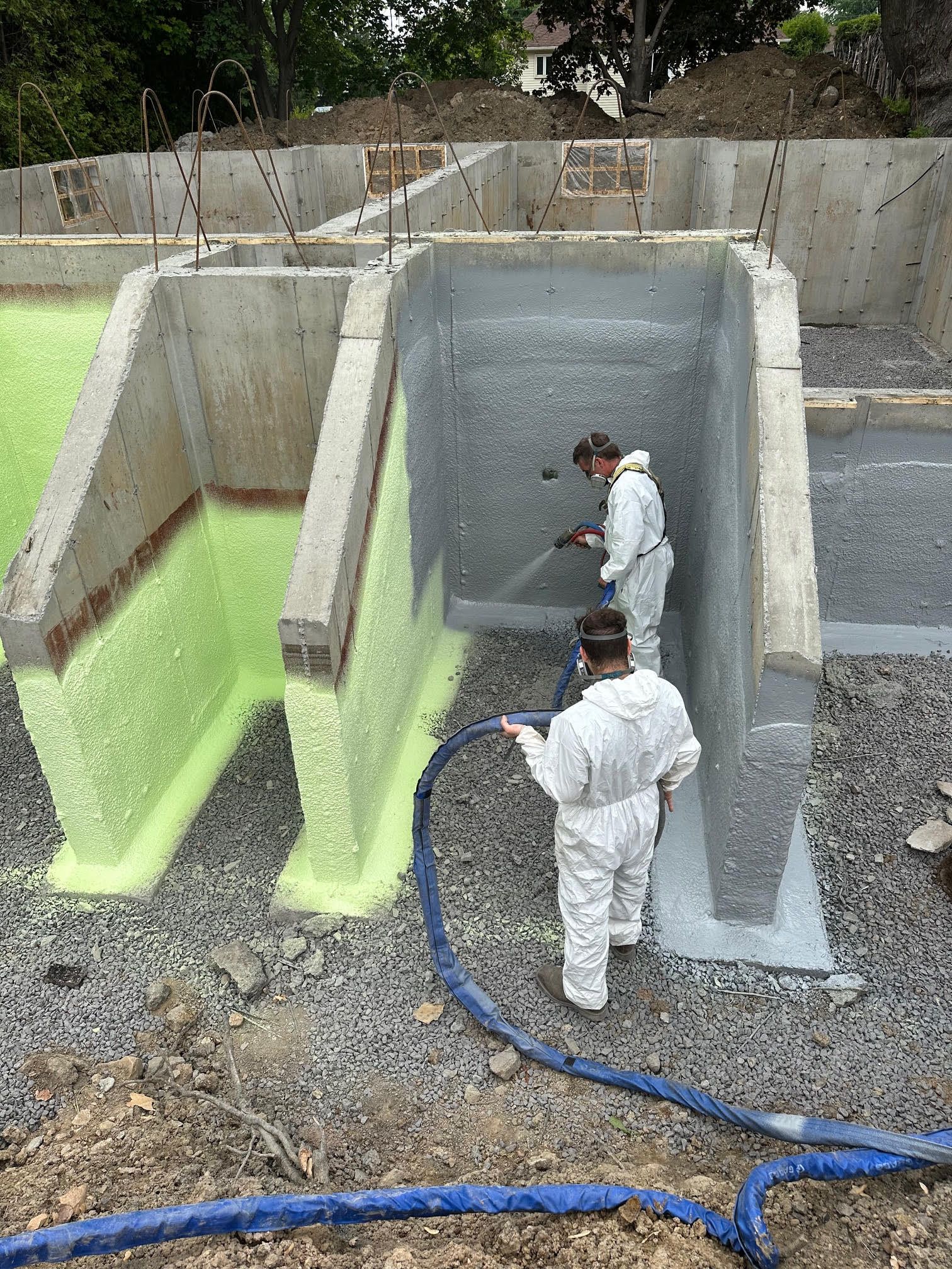 Deux ouvriers en combinaisons blanches pulvérisent de l'isolant sur des murs en béton sur un chantier de construction.