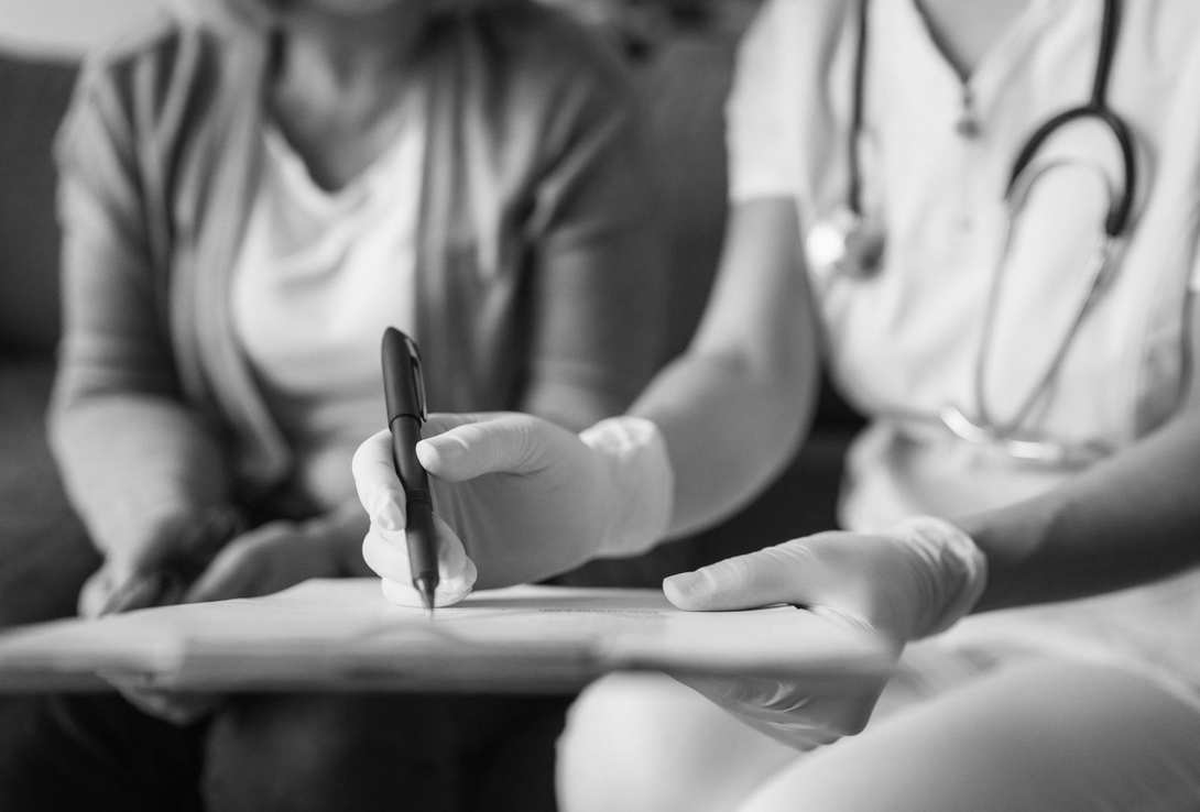Nurse writing on a clipboard while consulting with a patient; both are seated.