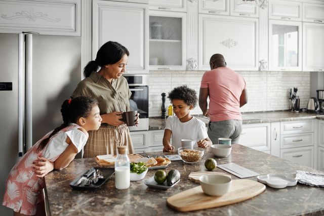 Family in kitchen, preparing breakfast. Mom with cup, kids eating cereal, dad cooking. Bright, sunny setting.