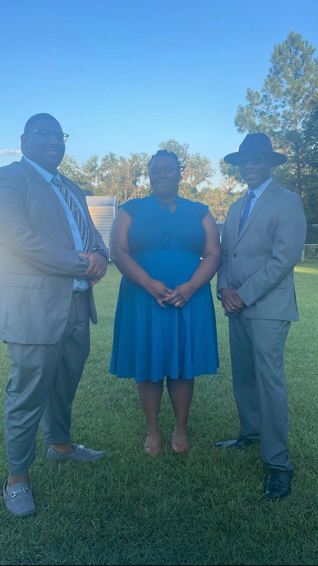Three people pose outdoors: one in a teal dress, two in suits. All are standing on grass.