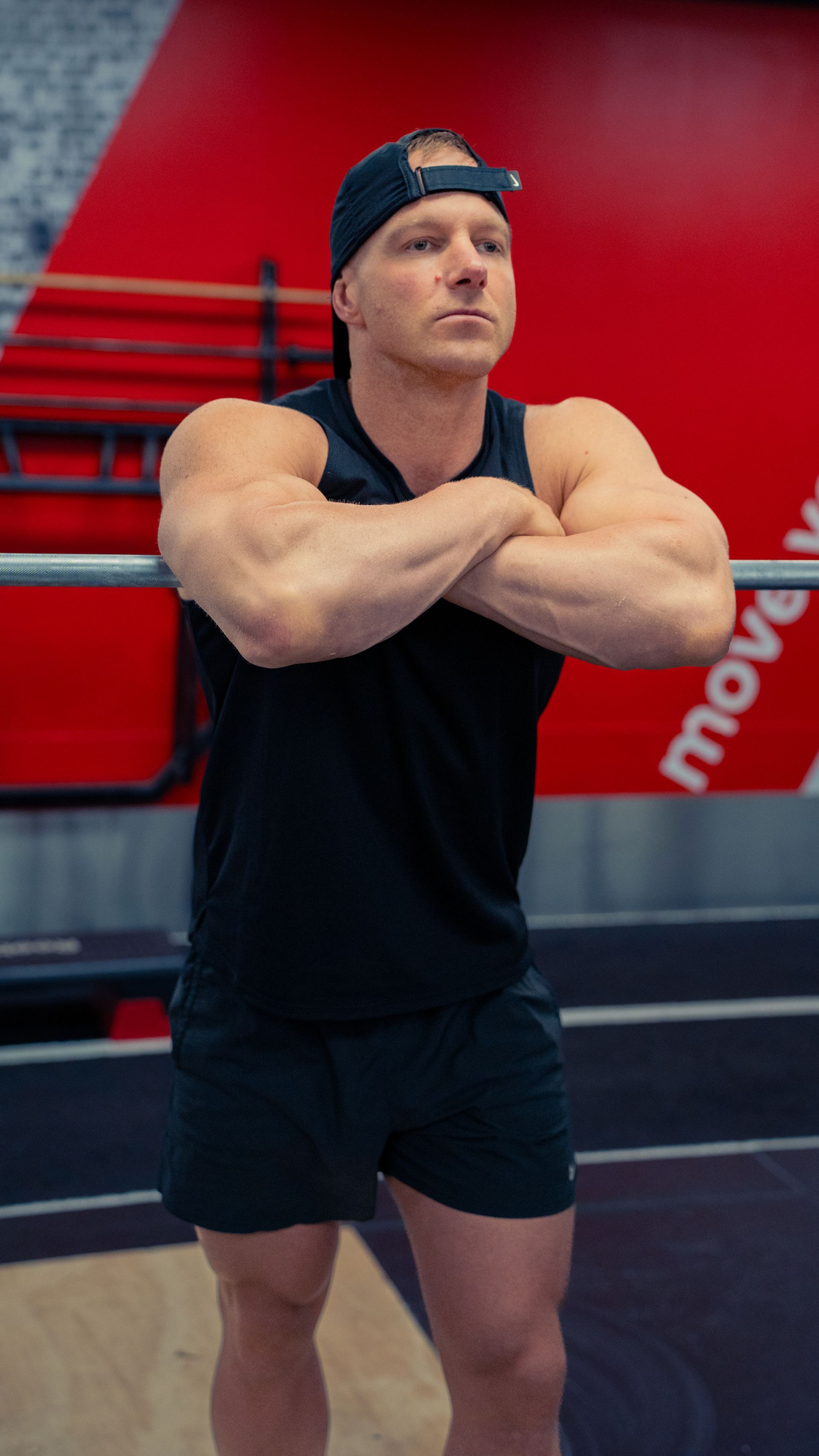 A man in a black tank top and shorts is standing with his arms crossed in a gym.