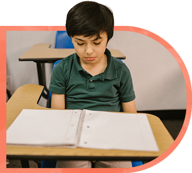 A young boy is sitting at a desk in a classroom reading a book.