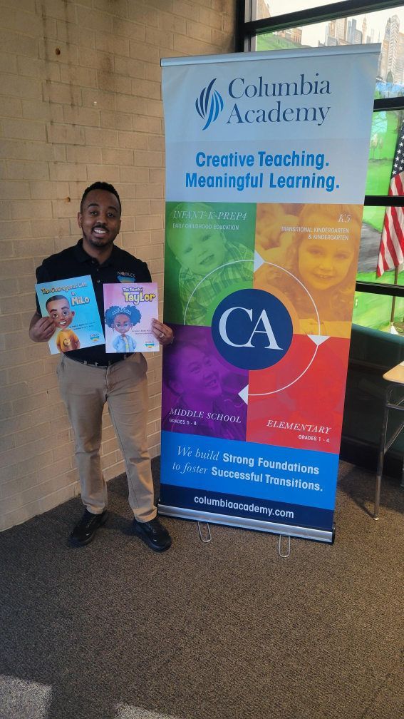 Myles Nobles stands smiling, holding his books next to a Columbia Academy promotional banner.