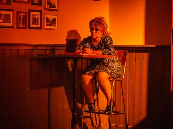 Woman seated at a bar in warm orange lighting, looking down at the table.