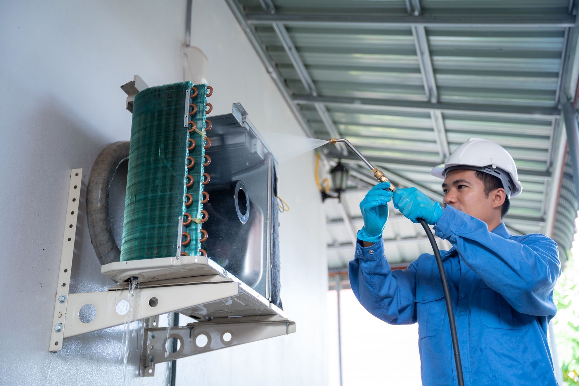 An HVAC technician in a blue jumpsuit cleans an outdoor AC unit with a spray nozzle.