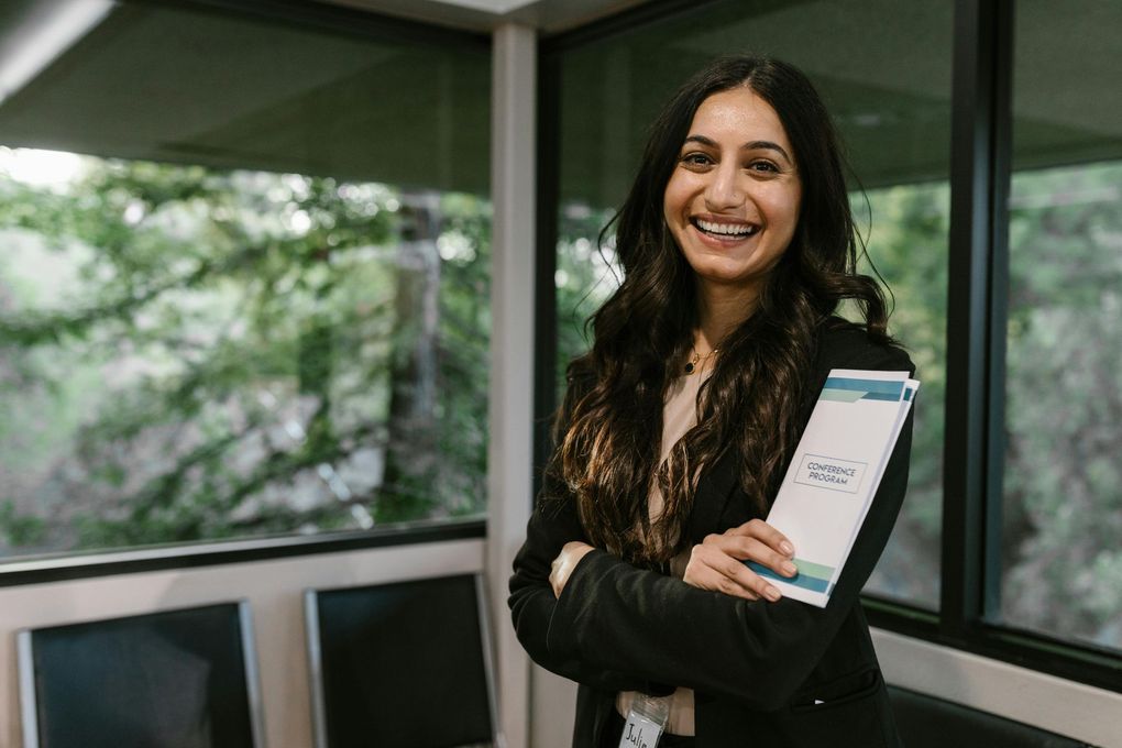 A woman is standing in front of a window holding a book and smiling.