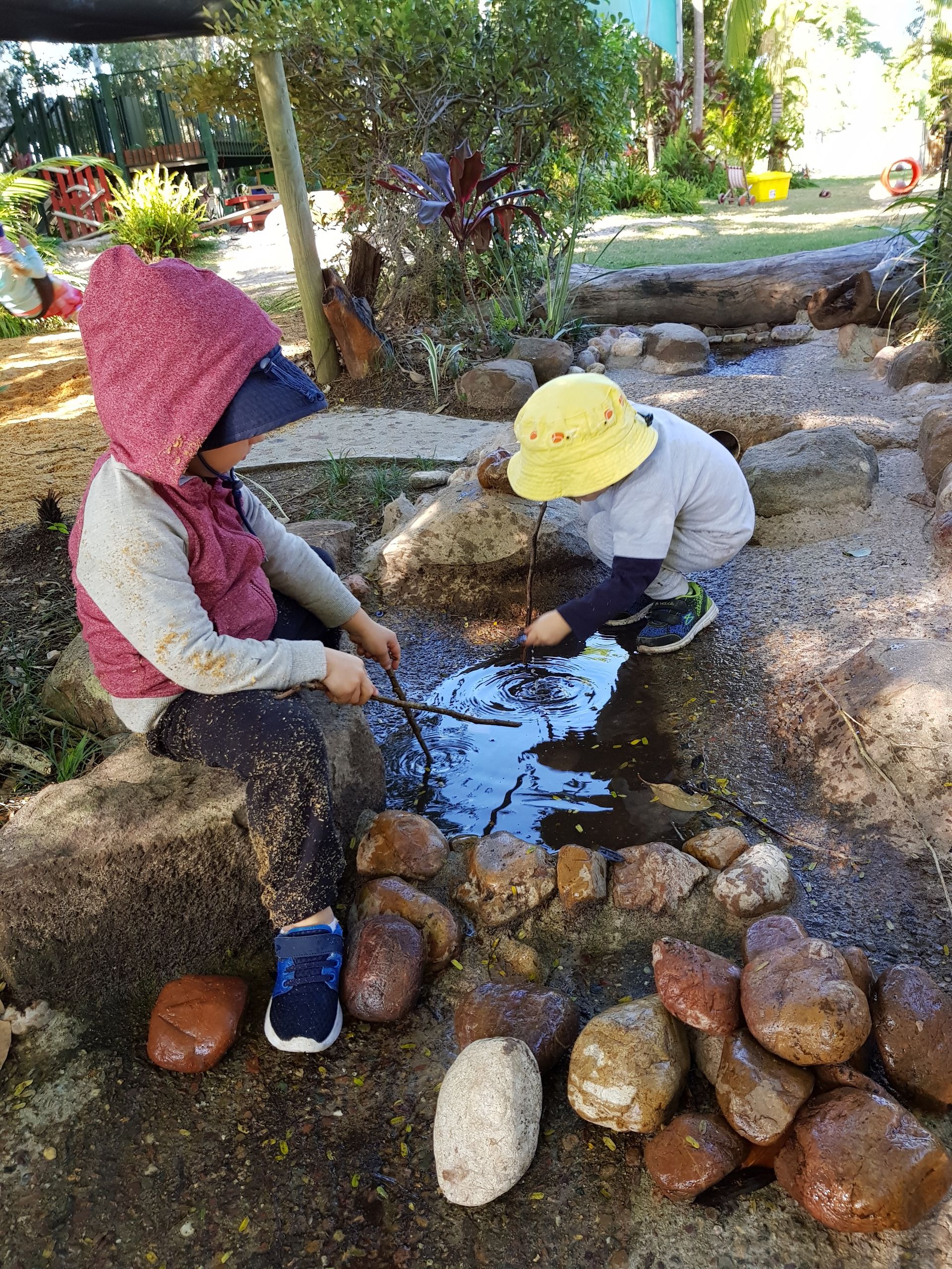 Two children playing by a small stream, one with a stick, the other looking into the water.