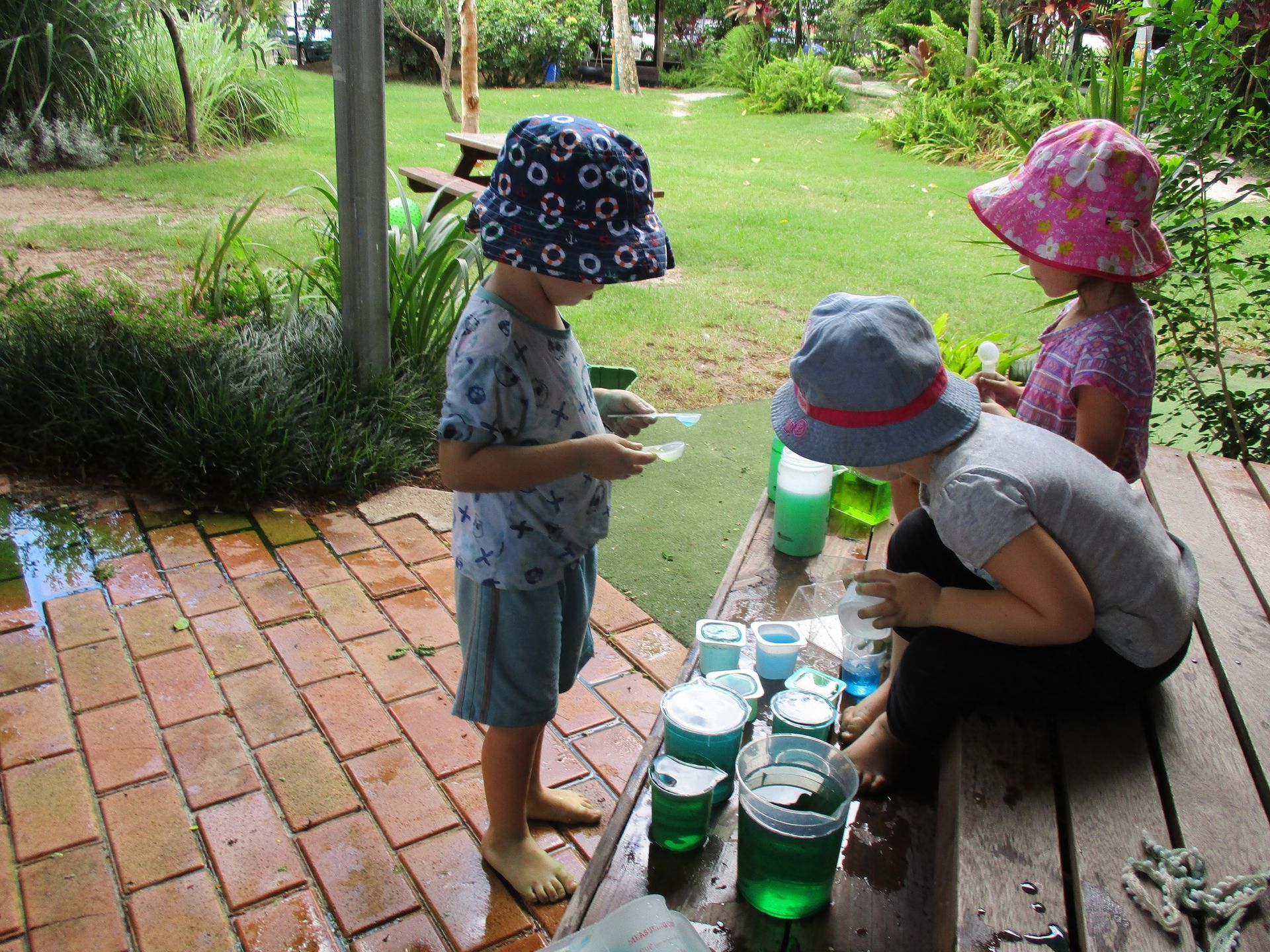 Children in hats experimenting with colorful liquids on a wooden table outdoors.