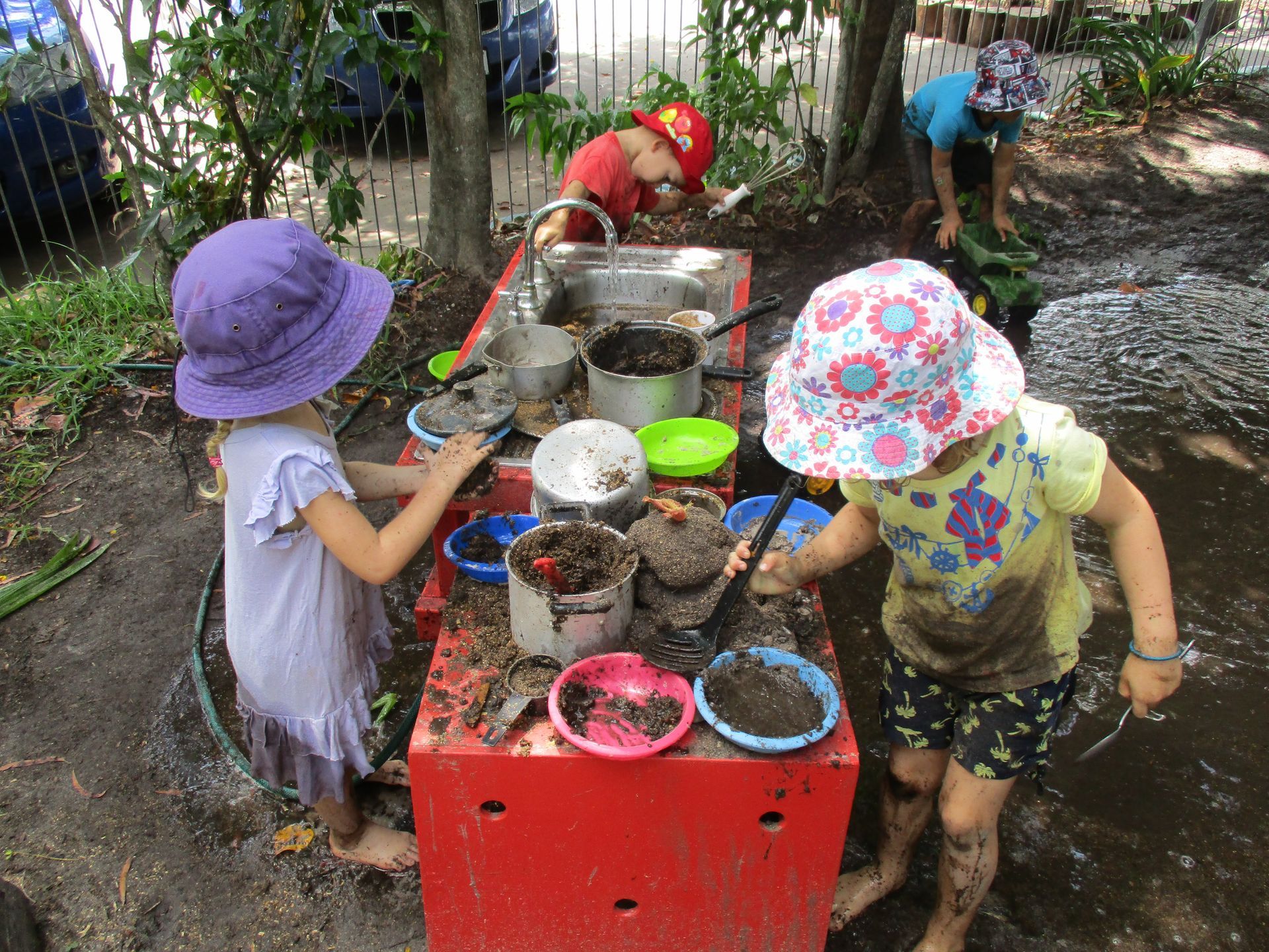 Two toddlers pull a wooden wagon down a path lined with green plants.