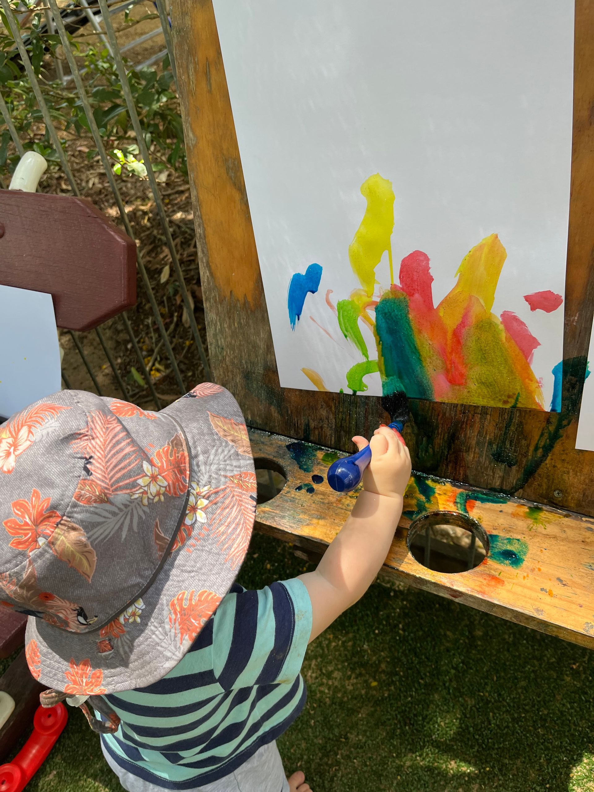 Child painting on a large easel with colorful paint outdoors; wearing a hat and striped shirt.