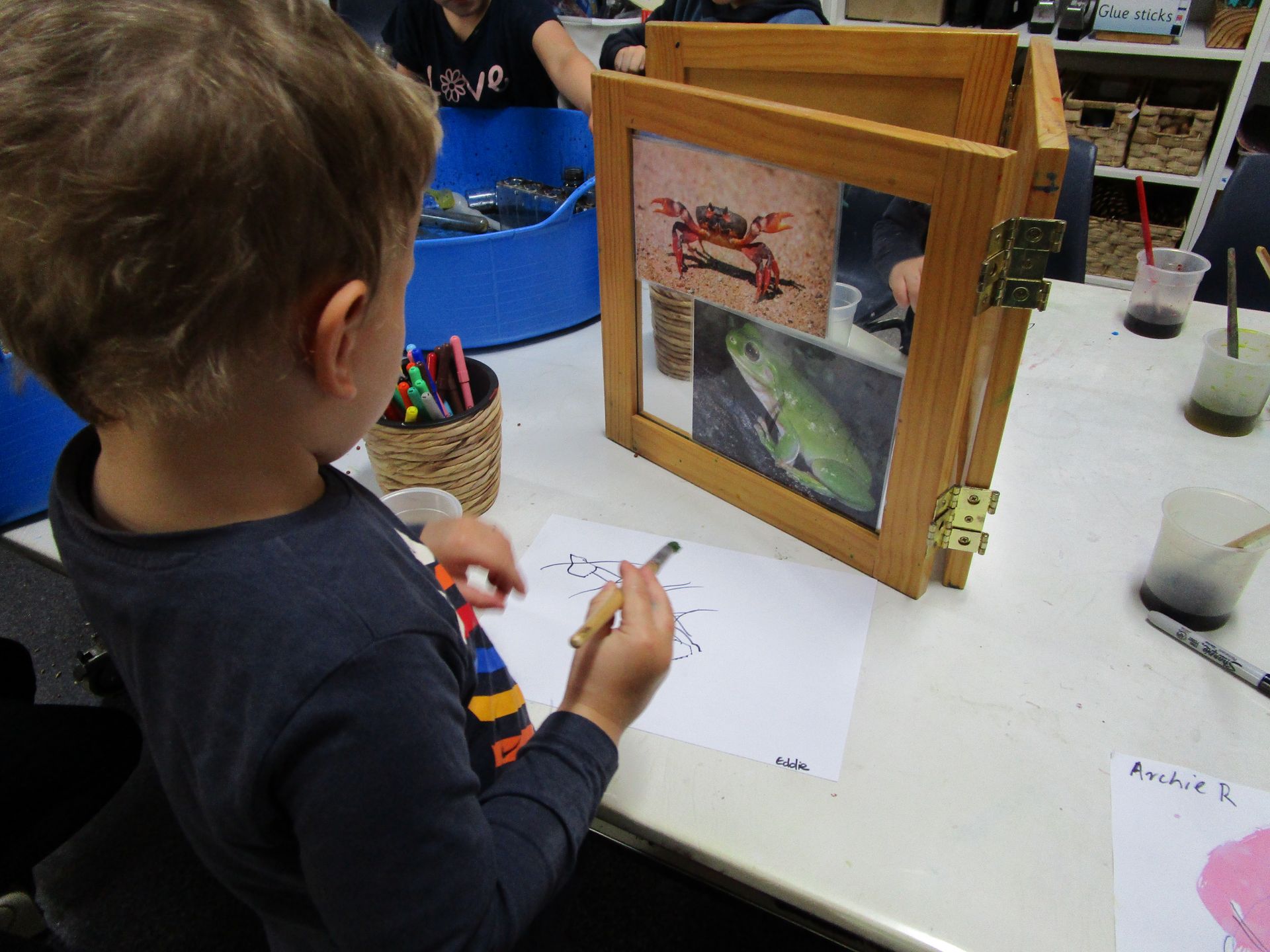 Child drawing at a table, looking at animal pictures in a wooden frame. Crayons and art supplies are nearby.