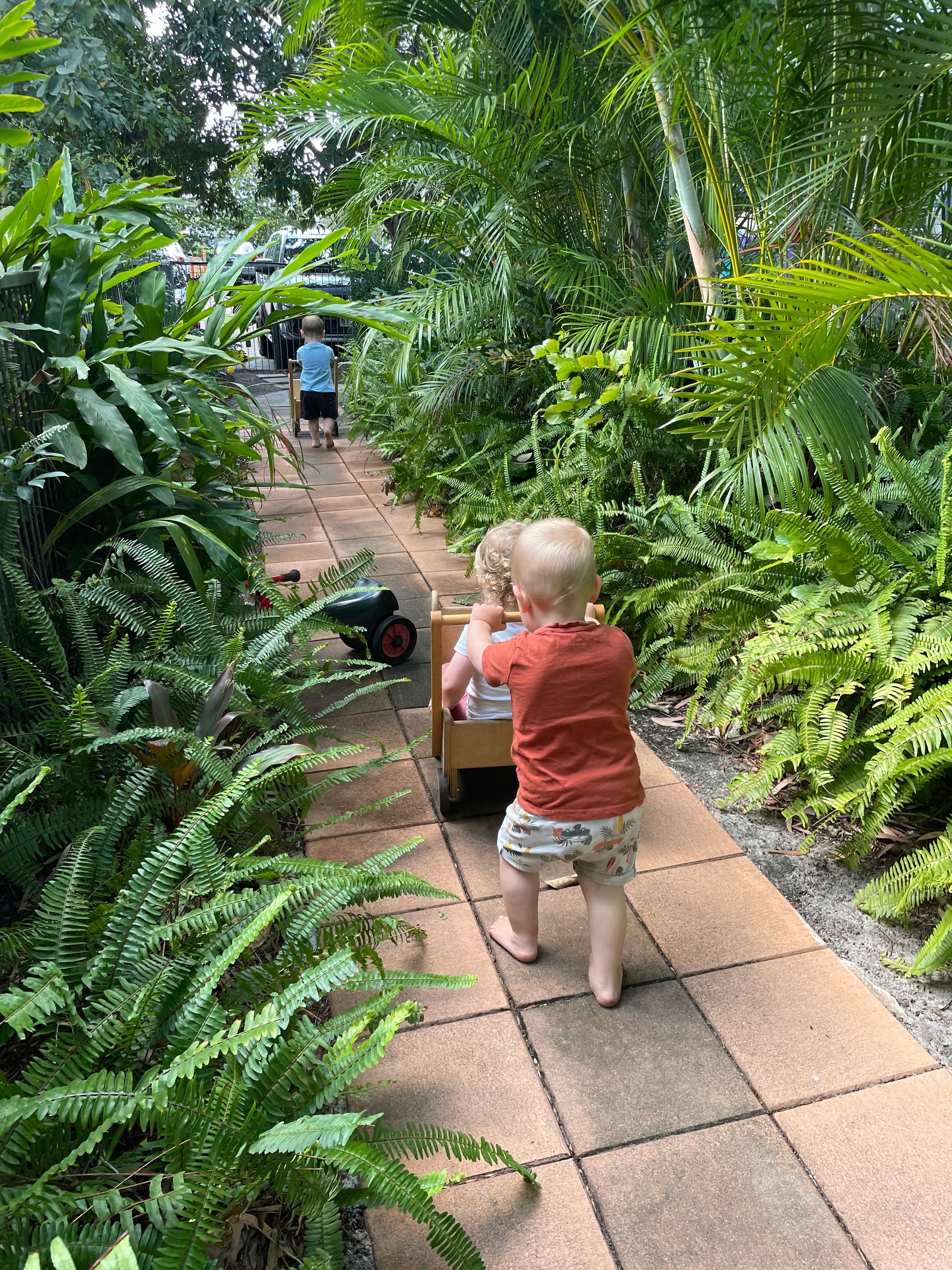 Two toddlers pulling a wagon down a stone path through lush greenery.