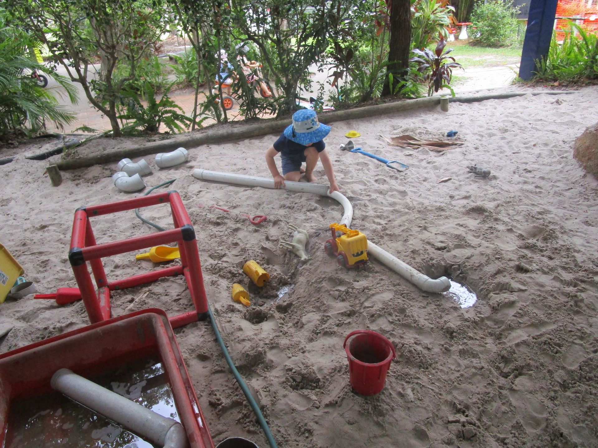 Child playing with sand and toys in a sandbox, using pipes and buckets.