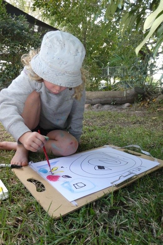 Young child with blonde hair painting outside on grass. They are wearing a hat and a sweater, and they are holding a paintbrush.