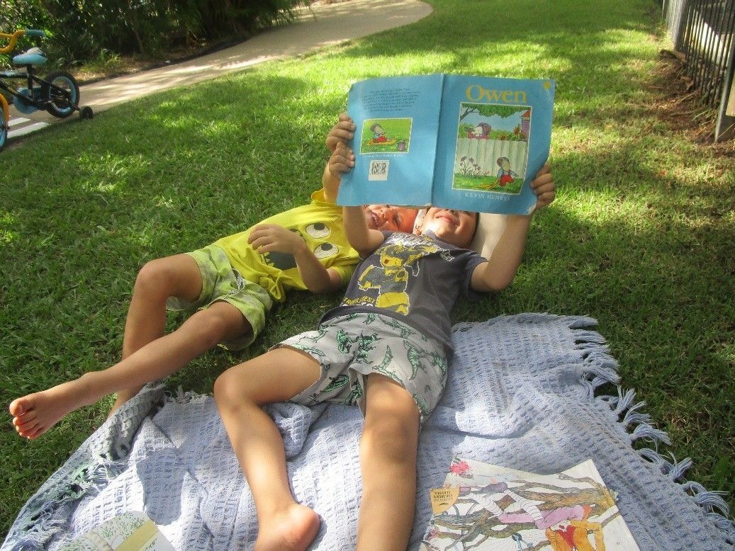 Two children lying on blanket, reading a book outdoors on green grass.