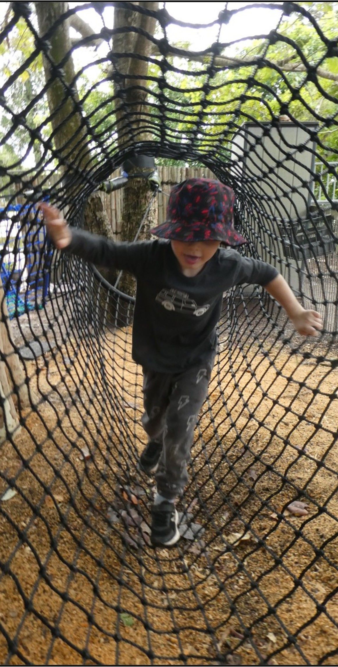 Boy in a hat walks through a netted tunnel, arms out for balance.