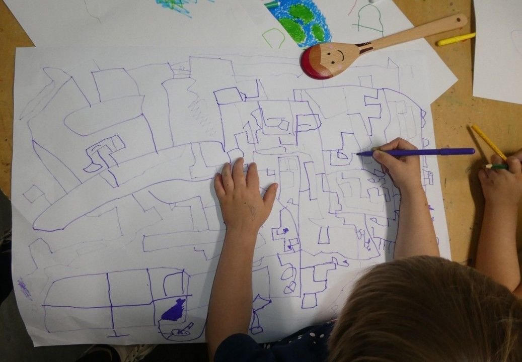 Child drawing with a purple pen on large paper, blue outline of buildings. Wooden spoon and crayons nearby.