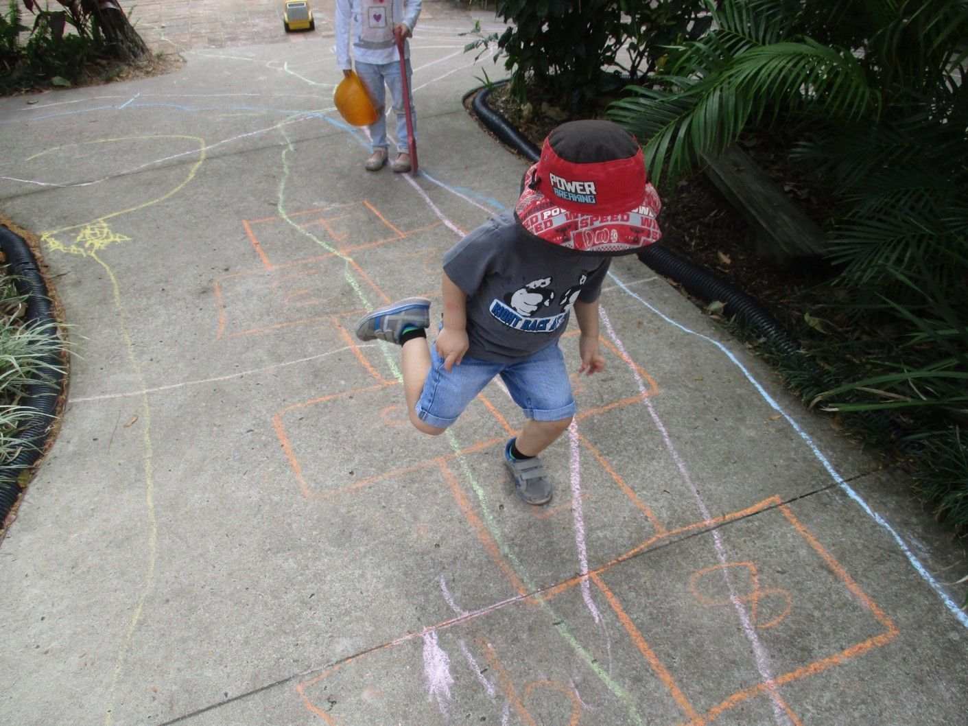 Child playing hopscotch on a concrete path, wearing a red hat and shorts, with another child in the background.
