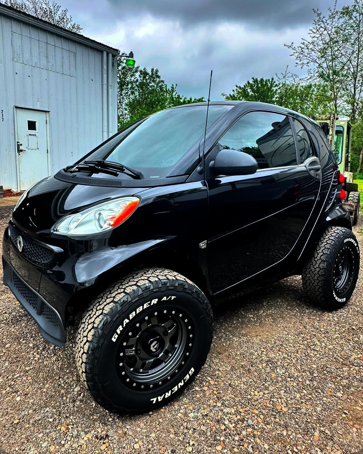 A black smart car is parked on gravel in front of a building.