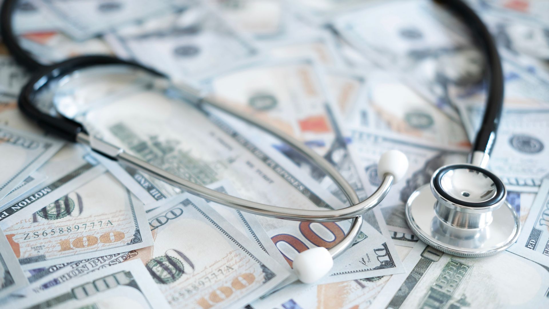 A silver medical stethoscope resting on top of a scattered pile of US one-hundred-dollar bills.