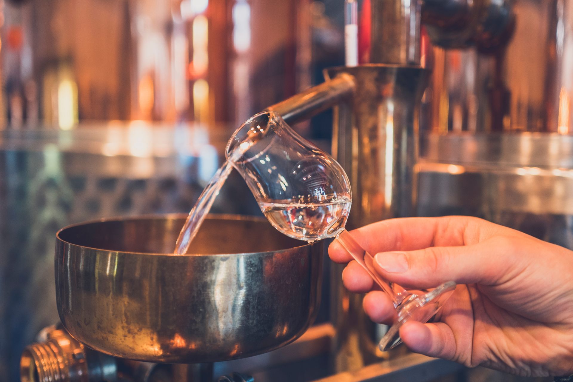 Hand pouring clear liquid from a glass into a dark cooking pot in a kitchen-like setting