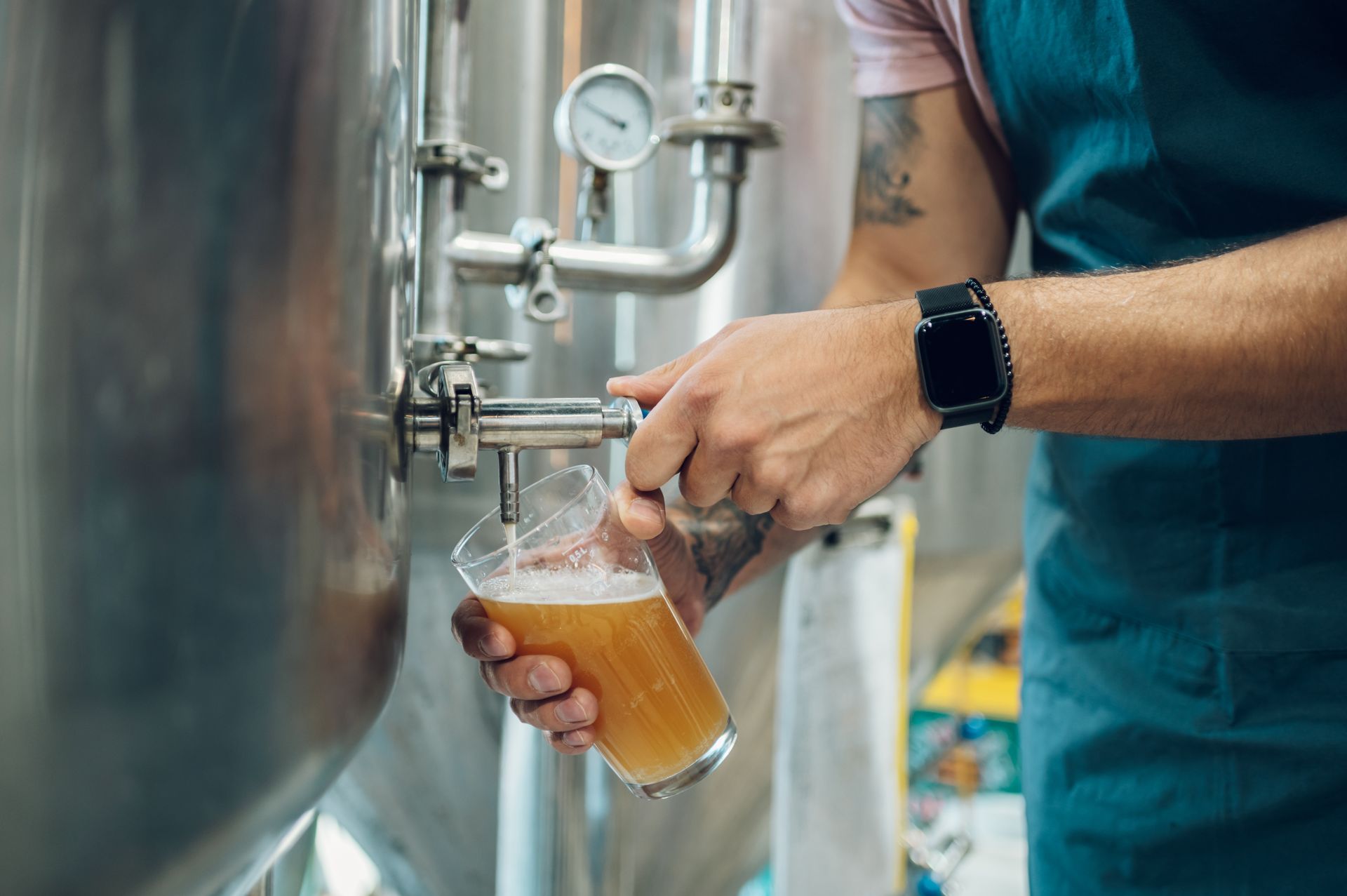 Person pouring beer from a tap into a plastic cup beside stainless steel tanks