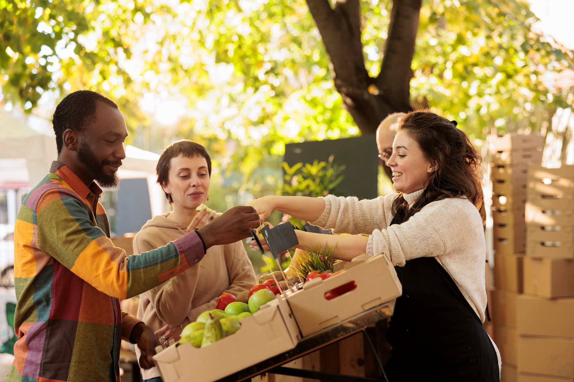 Three people exchange fresh produce at an outdoor market stall under sunny trees.