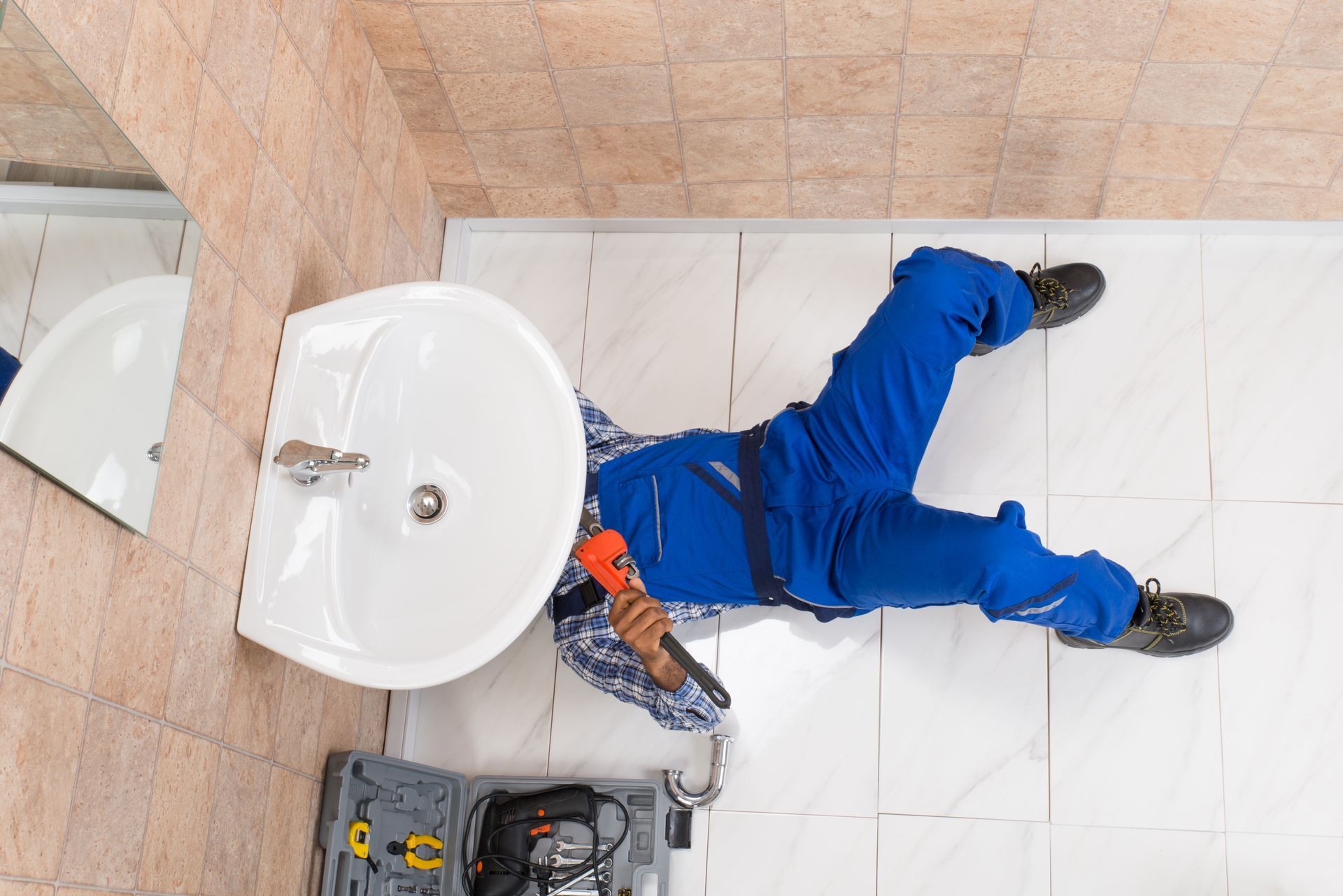 Plumber repairing bathroom sink with tools under vanity on hex tile floor during home plumbing job.