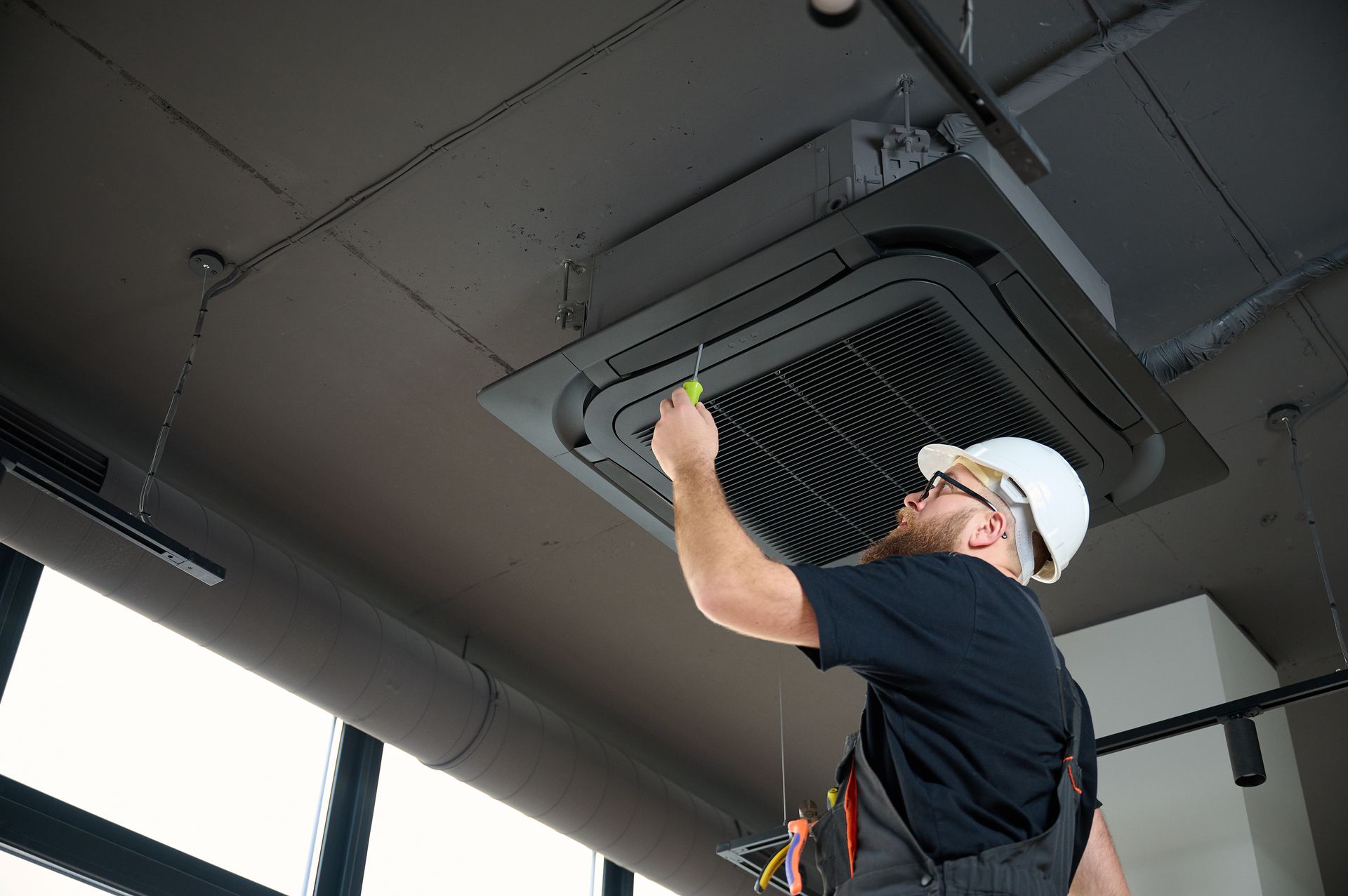 HVAC technician in white hardhat using a screwdriver on a ceiling-mounted air conditioning unit.