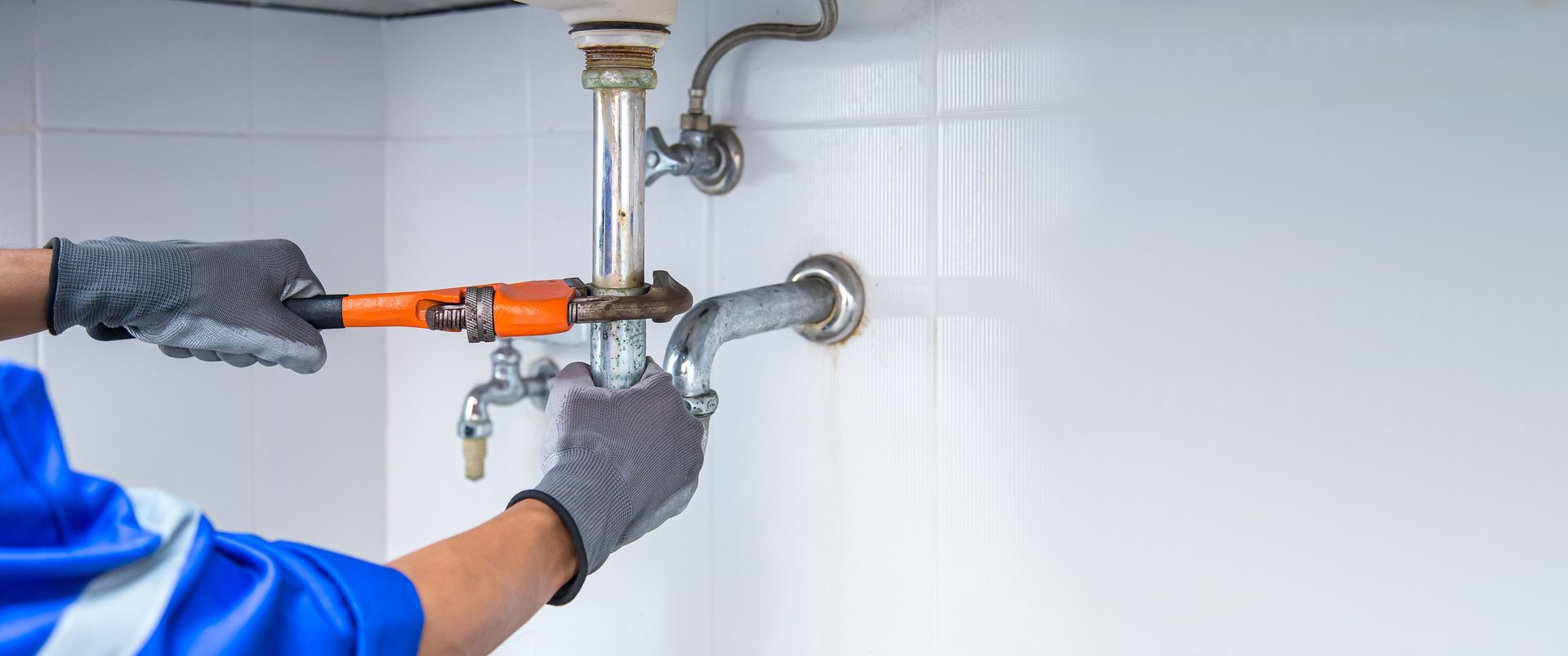 Gloved hands using a pipe wrench to tighten plumbing under a sink with visible metal pipes.