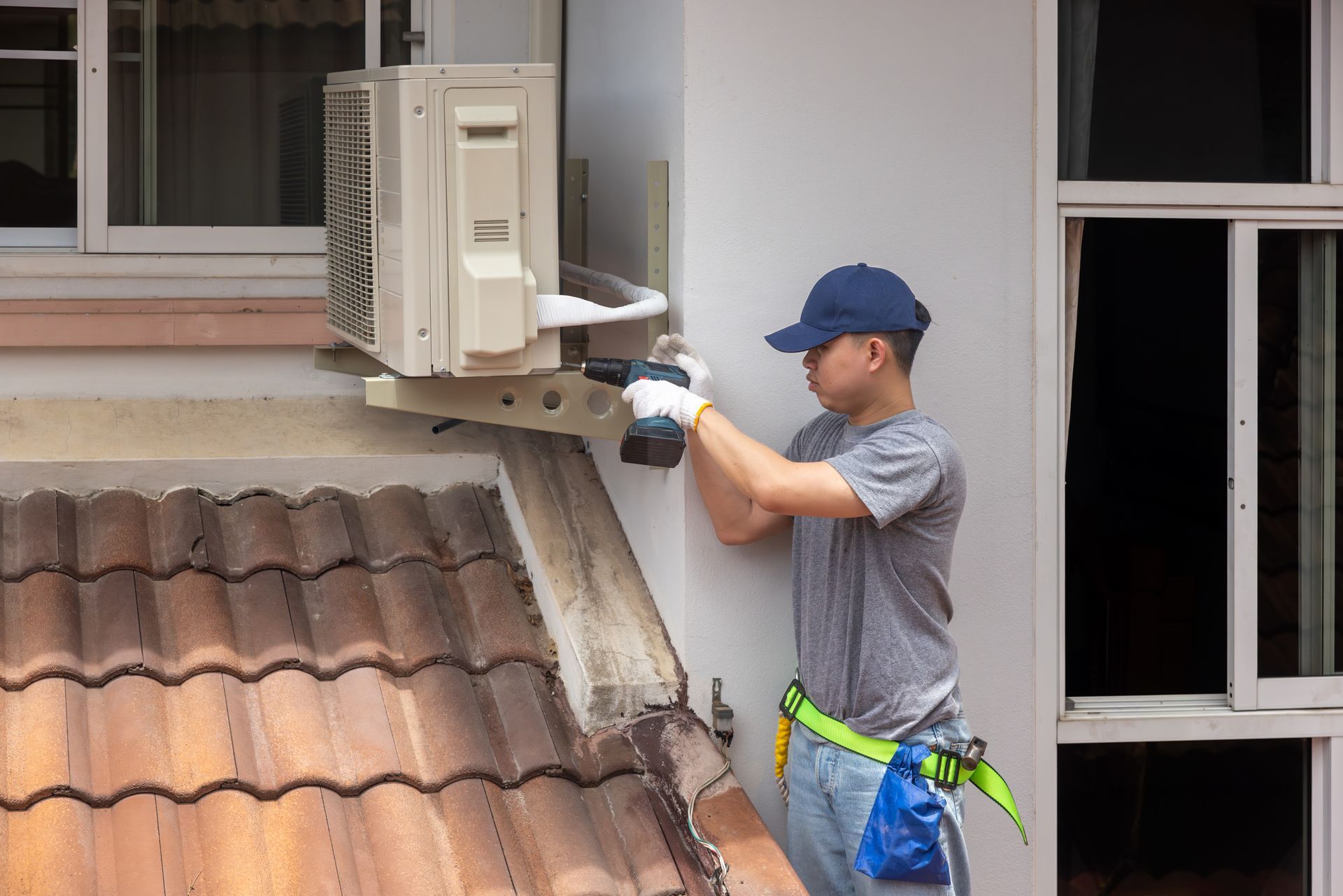 Technician installing or repairing a wall-mounted air conditioner unit beside a window on a tiled roof. Technician installing or repairing a wall-mounted air conditioner unit beside a window on a tiled roof.