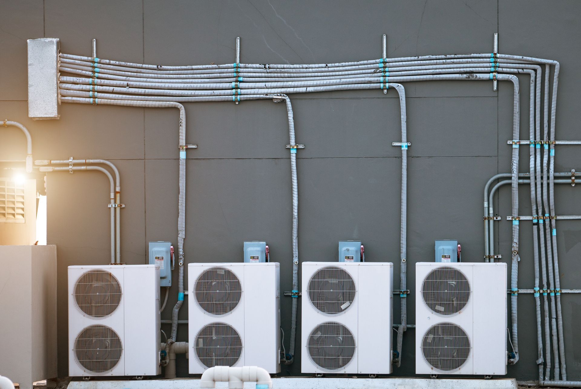 Four white air conditioning units mounted on a grey wall with conduits.