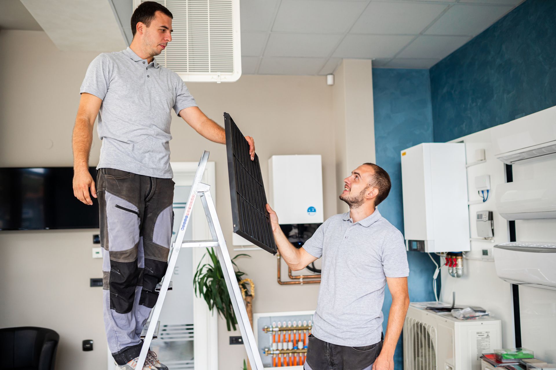 Two commercial AC contractors, one on a ladder, are holding parts in an office space.