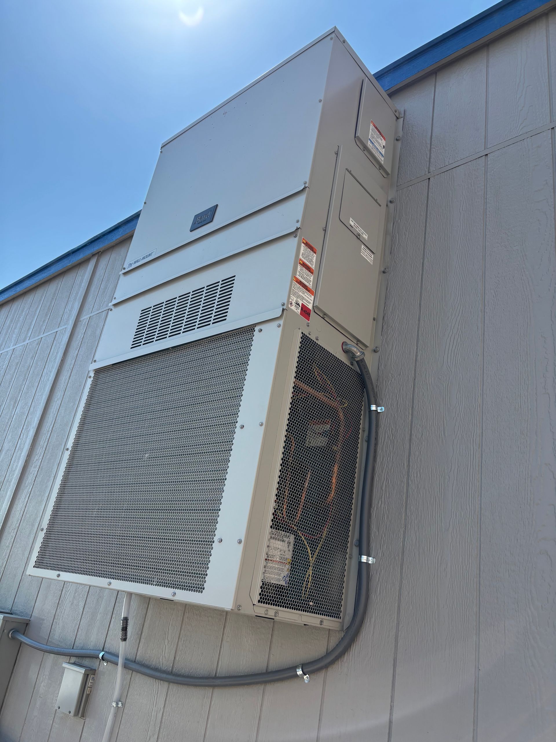 Air conditioner mounted on a light gray building, with a silver metal exterior and a gray conduit.